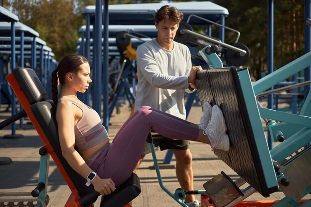 Młoda kobieta ćwicząca na maszynie do przywodzenia ud, młody mężczyzna pomagający